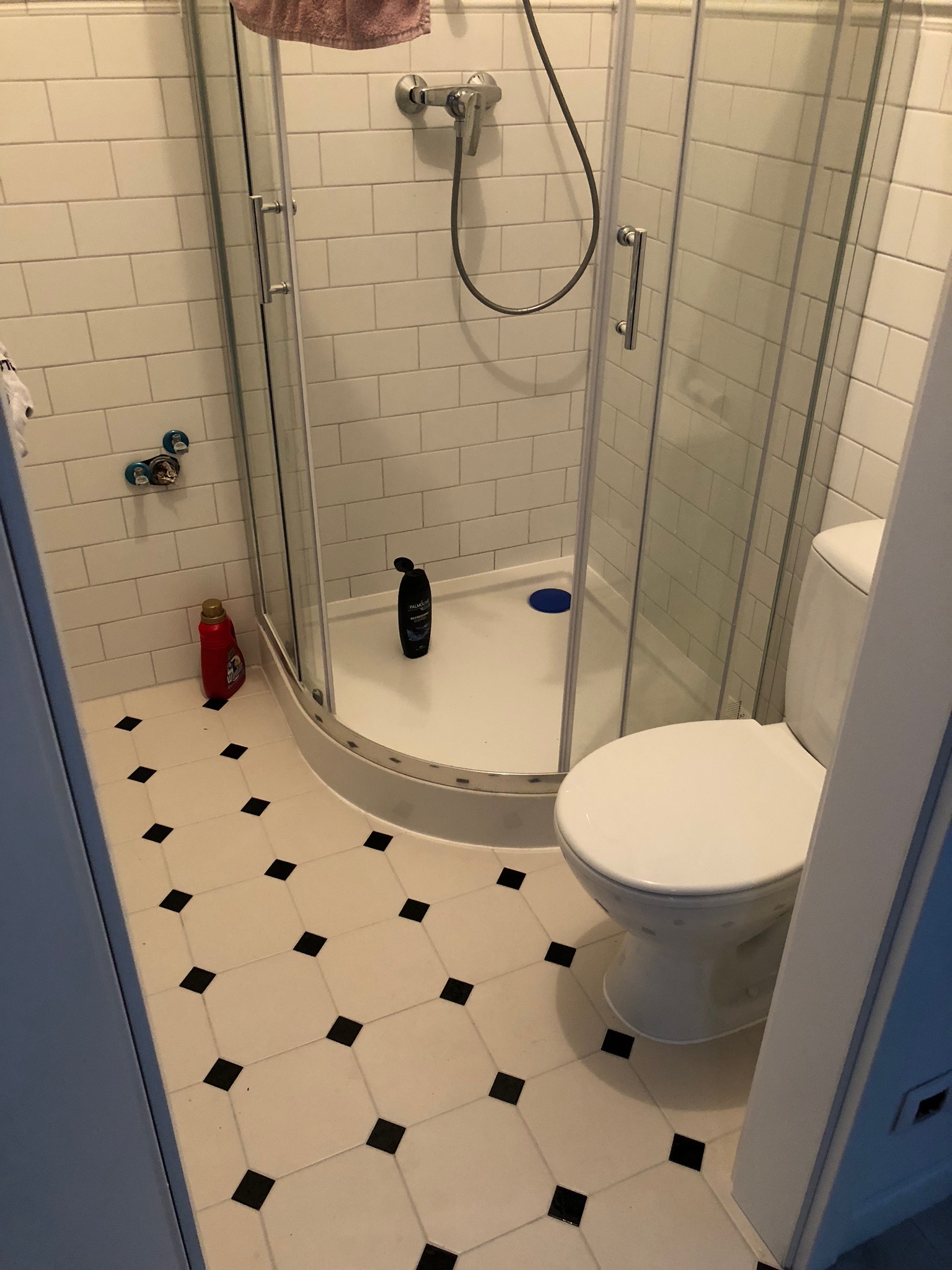 Ujęcie łazienki z kabiną prysznicową z zaokrąglonymi drzwiami, toaletą, białą glazurą na ścianach i podłogą w biało-czarną szachownicę; widoczna butelka płynu do prania obok kabiny.