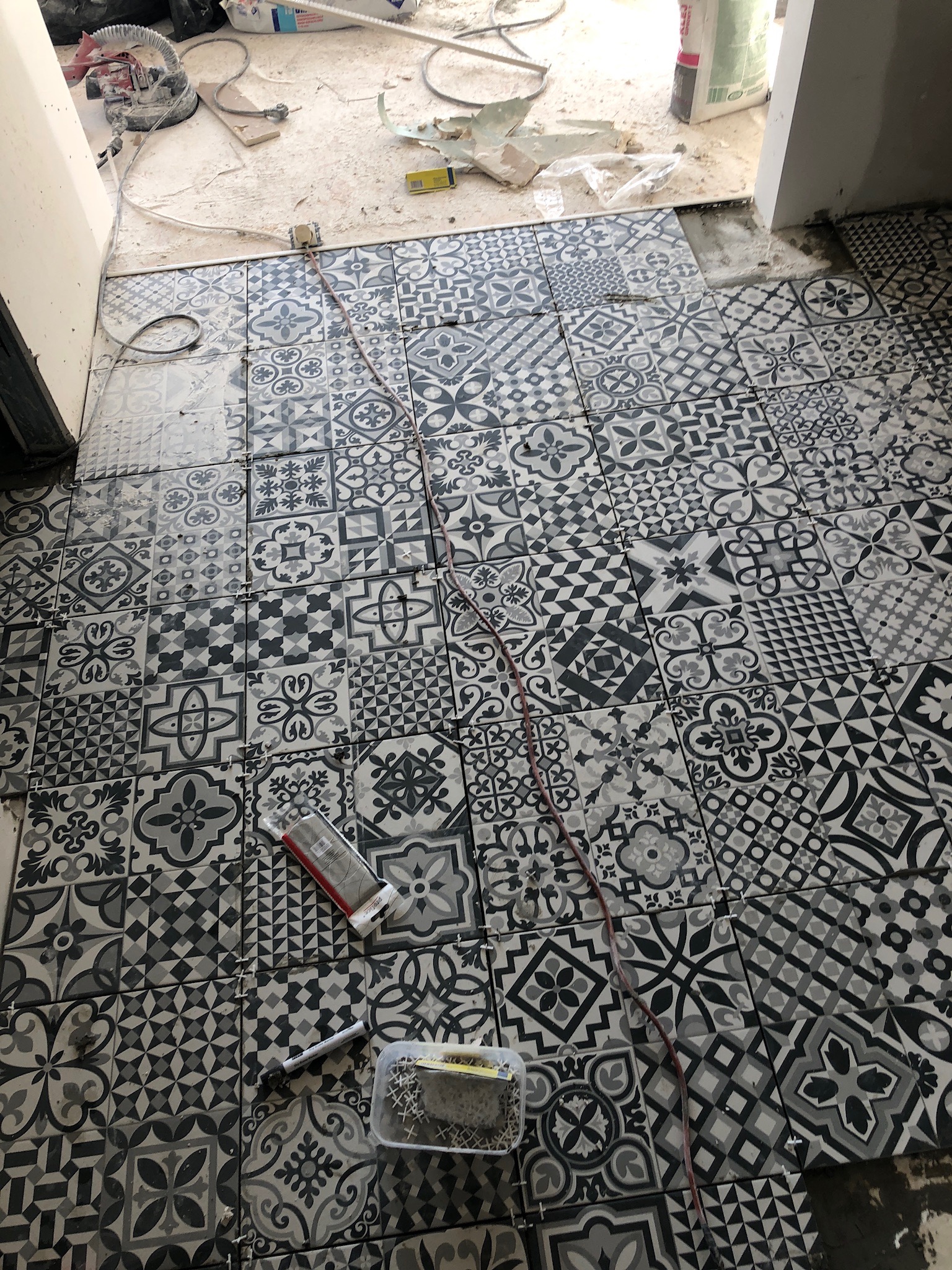 Układanie wzorzystych, czarno-białych płytek podłogowych w trakcie prac, widoczne krzyżyki dystansowe, opakowanie kleju i przewód elektryczny na powierzchni.