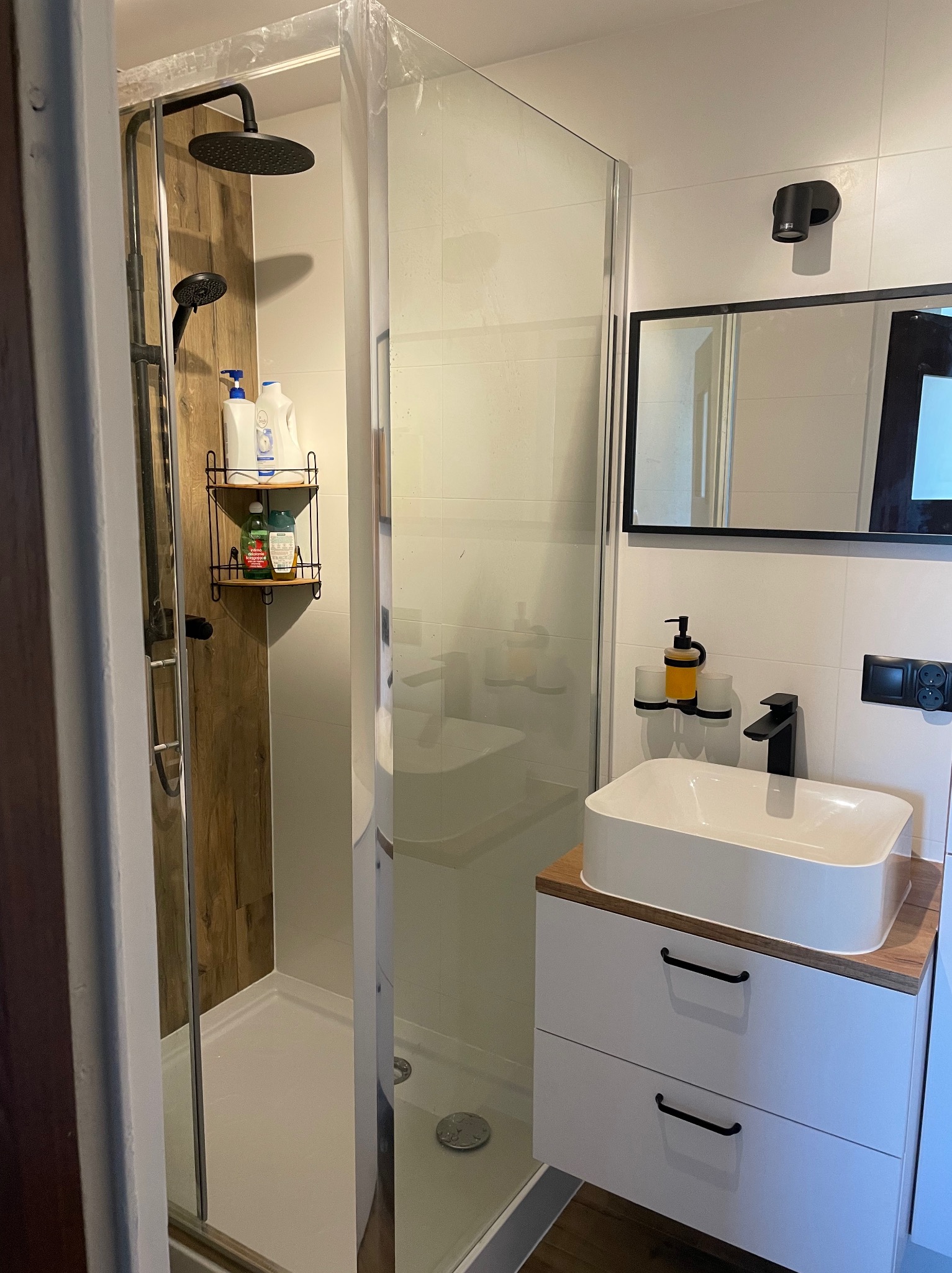 Nowoczesna łazienka z kabiną prysznicową typu walk-in, umywalką nablatową na szafce z dwiema szufladami, czarną armaturą i drewnianymi akcentami.