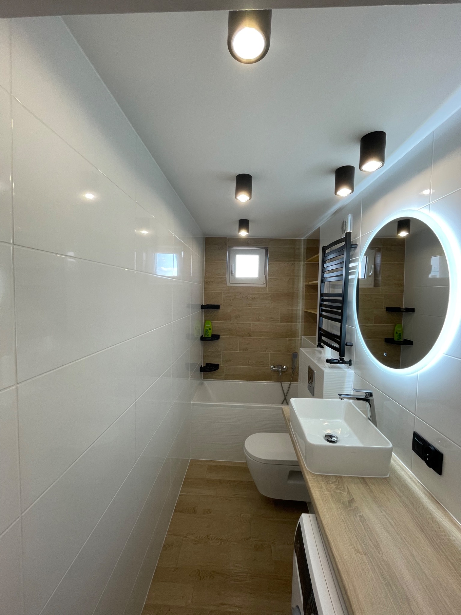 Nowoczesna łazienka po remoncie: białe płytki, drewnopodobna podłoga, okrągłe lustro z podświetleniem LED, minimalistyczna umywalka nablatowa na blacie z drewnopodobnego laminatu, czarne lampy...