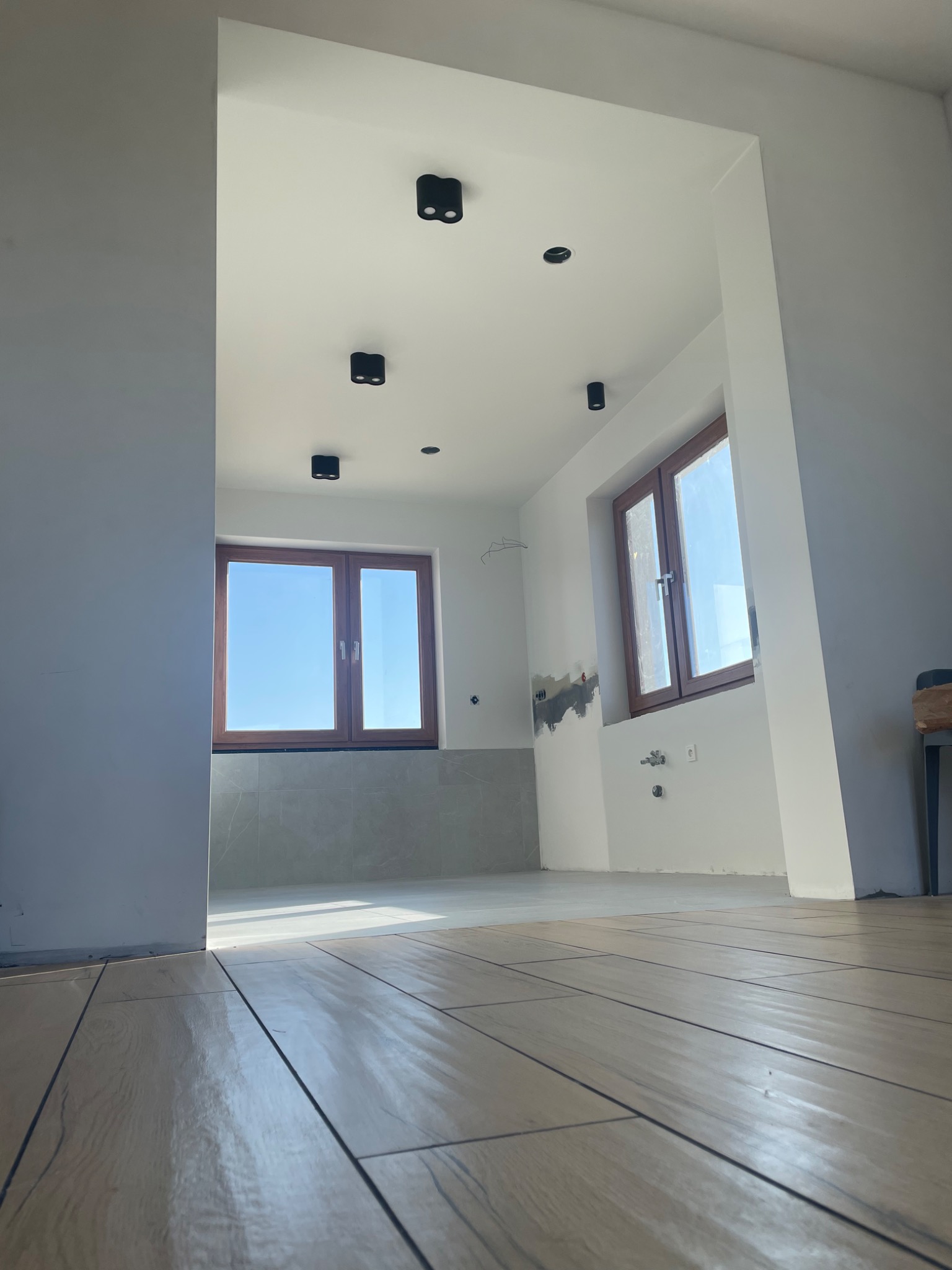 Wnętrze w trakcie remontu: ułożone płytki podłogowe imitujące drewno, częściowo wykończona ściana z szarą glazurą, przygotowane punkty świetlne na suficie, widoczne instalacje hydrauliczne na ścianie.