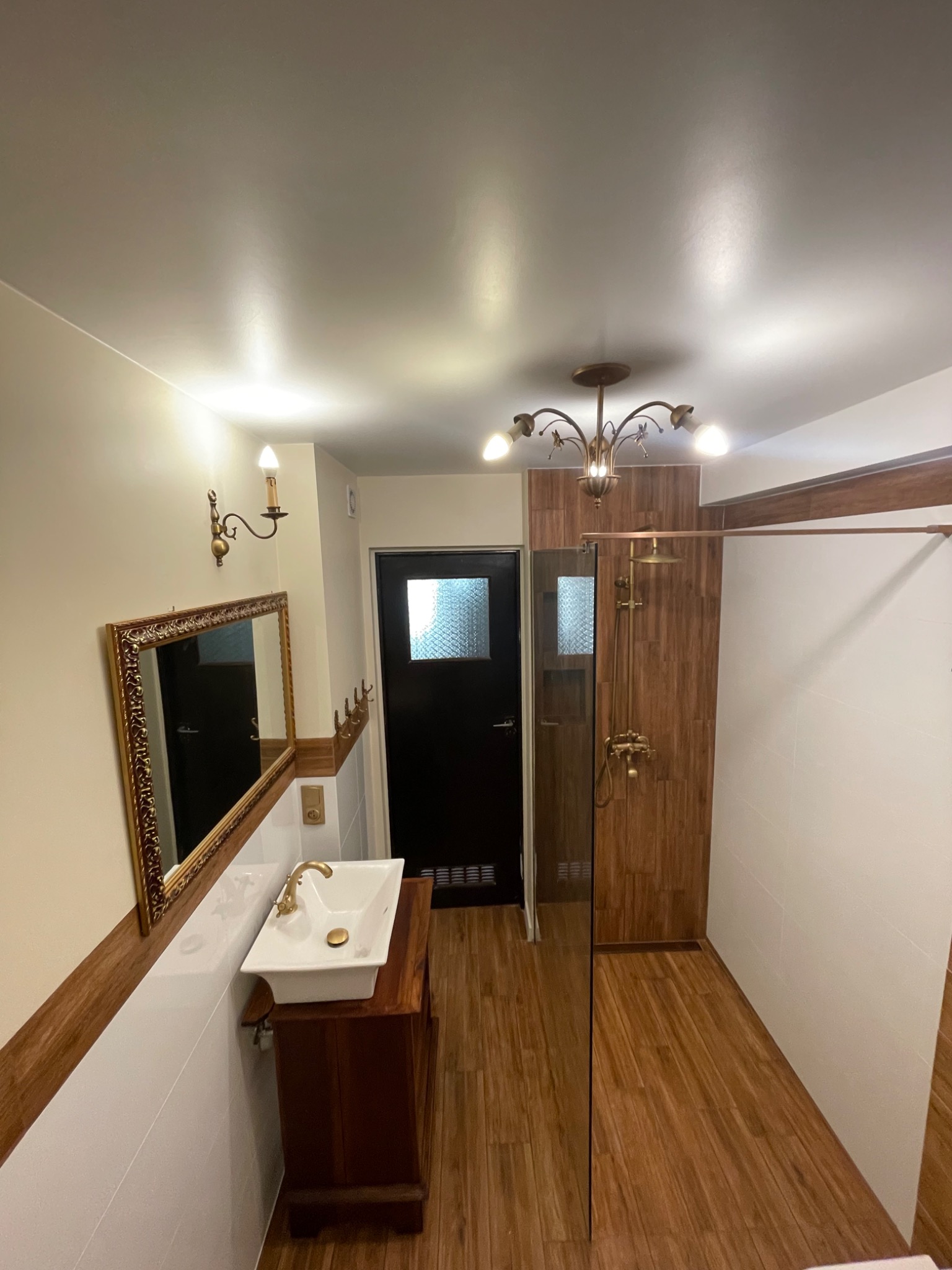 Wykończona łazienka w stylu retro z drewnianą podłogą, kabiną prysznicową z panelami imitującymi drewno, umywalką na drewnianej szafce i złotymi detalami oraz lustrem w ozdobnej ramie.