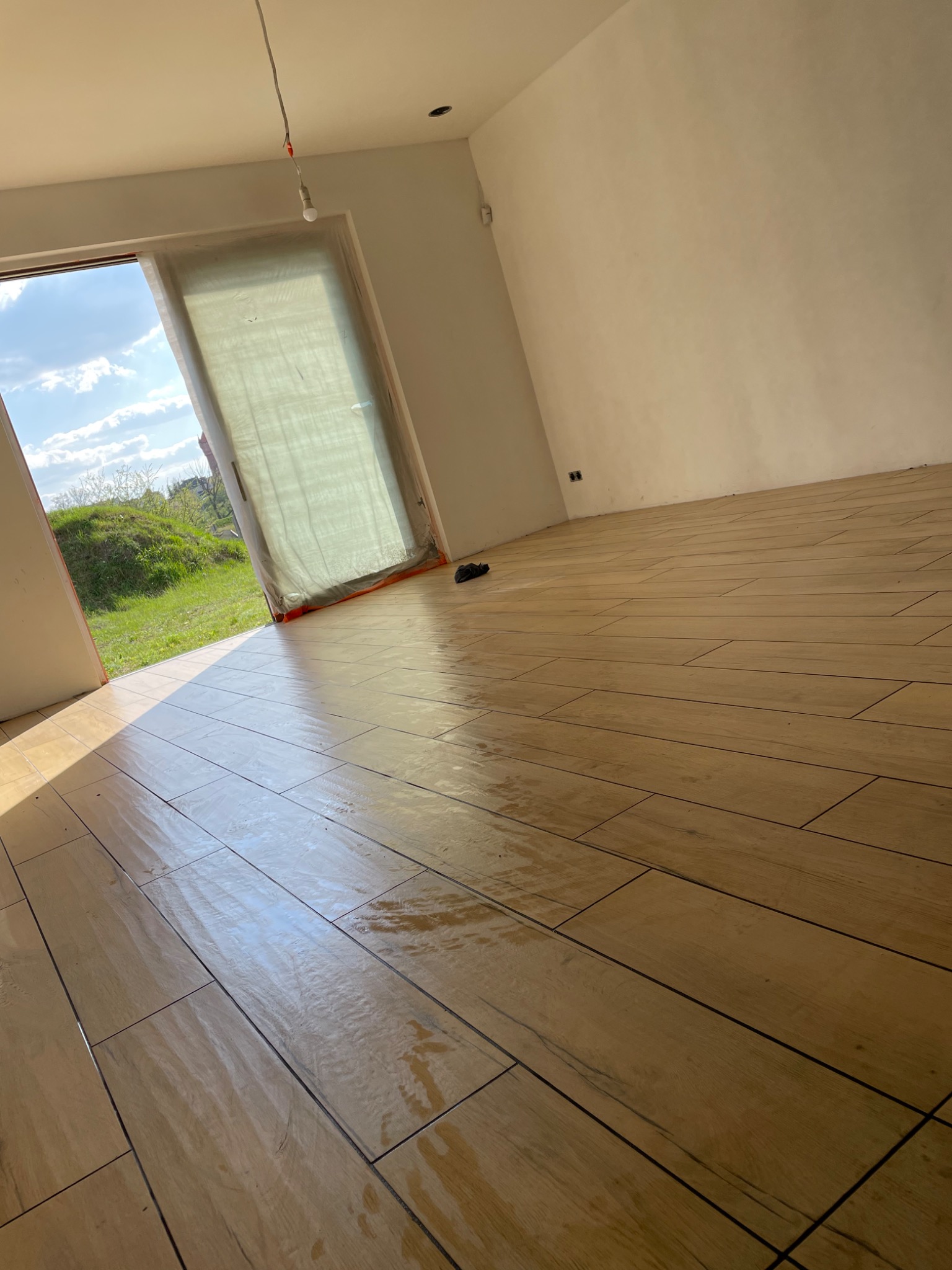 Ułożona podłoga z płytek imitujących drewno w jasnym, pustym pokoju z widokiem na zielony krajobraz przez otwarte drzwi balkonowe, częściowo zasłonięte folią ochronną.