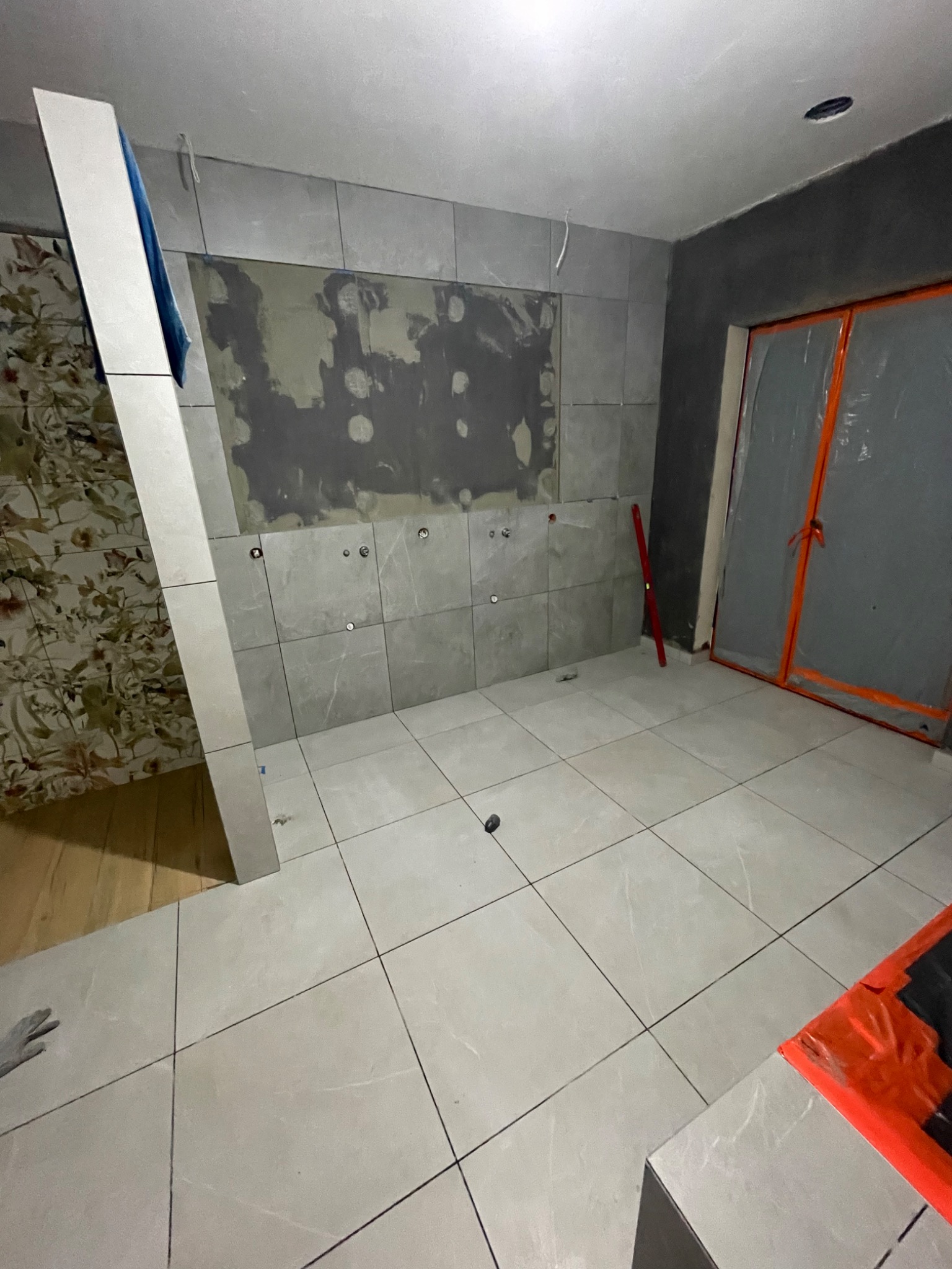 Wnętrze łazienki w trakcie układania płytek ceramicznych na ścianach i podłodze, widoczne przygotowane otwory na armaturę, fragment ściany z tapetą i drzwi zabezpieczone folią ochronną.