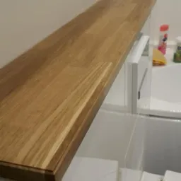 Drewniany blat zamontowany nad białą zabudową w łazience, widoczna tekstura drewna i fragment instalacji wodnej w tle.