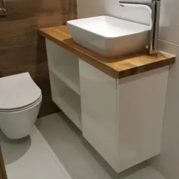 Nowoczesna łazienka z białą szafką pod umywalkę z drewnianym blatem, kwadratową umywalką nablatową i minimalistyczną baterią, obok toaleta.