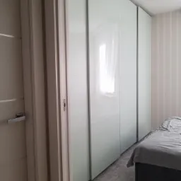 Biała szafa przesuwna z lustrzanymi drzwiami w sypialni z tapetą w pionowe paski, widoczny fragment łóżka z szarą pościelą.