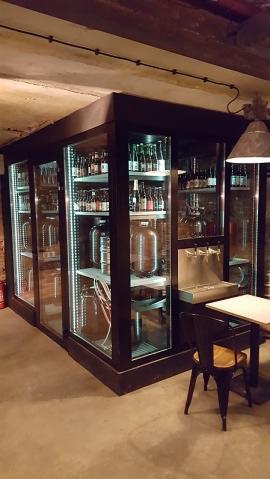 Duża, przeszklona szafa chłodnicza z ciemną ramą, zawierająca butelki piwa, metalowe beczki i nalewaki, ustawiona w lokalu z surowym betonowym sufitem i podłogą.