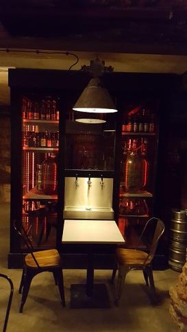 Loftowa aranżacja z ciemną szafą ekspozycyjną podświetloną czerwonym światłem, metalowym oświetleniem industrialnym i barem z trzema kranami oraz dwoma krzesłami o drewnianych siedziskach.