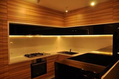 Nowoczesna kuchnia z drewnianymi frontami szafek, białym blatem i podświetleniem LED pod szafkami wiszącymi, widoczna płyta grzewcza i piekarnik w zabudowie.