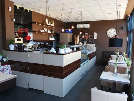 Wnętrze kawiarni z ladą barową wykończoną w odcieniach bieli i ciemnego drewna, wiszącymi lampami, stolikami i krzesłami, oraz dekoracyjnym zegarem na ścianie.