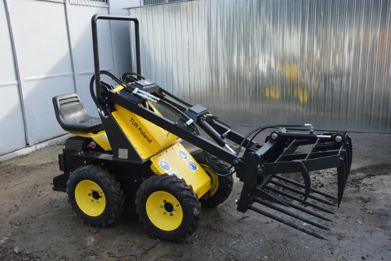 Żółto-czarna miniładowarka TUR Poland z widłami do obornika na błotnistym podłożu, z blachy falistej w tle.