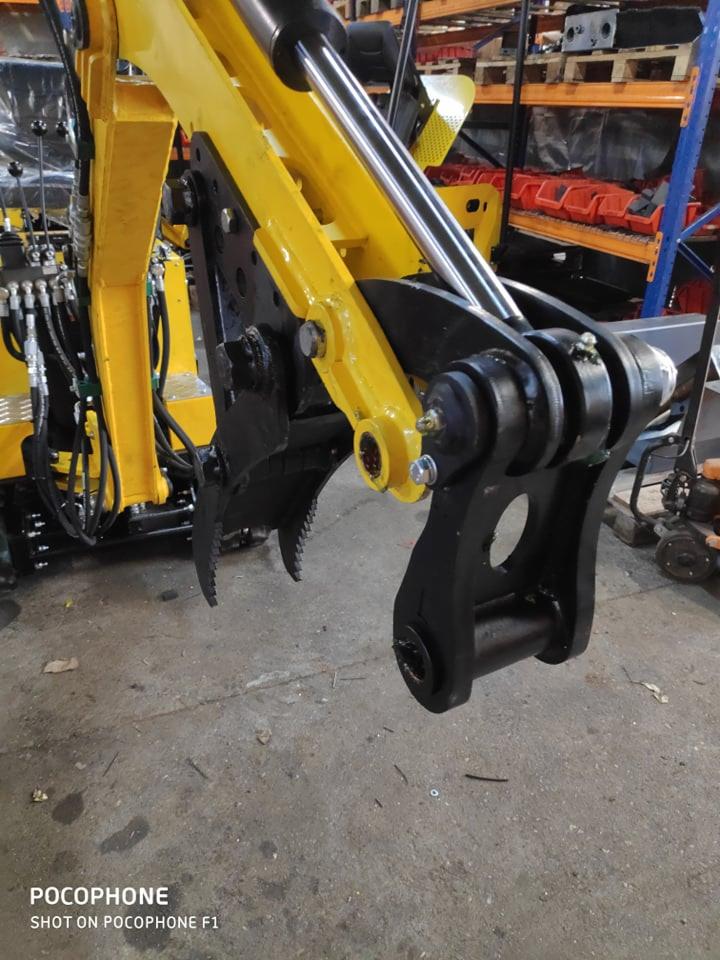 Żółto-czarna maszyna budowlana z hydraulicznym ramieniem i chwytakiem, widoczne przewody hydrauliczne i metalowe regały w tle.