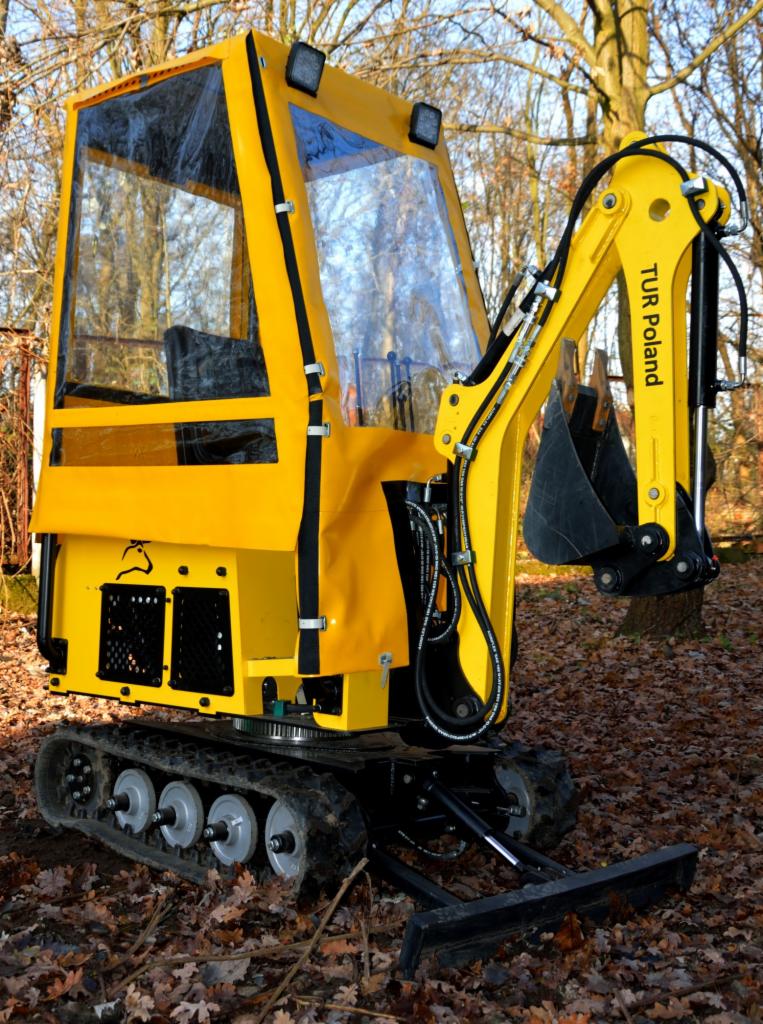 Żółta, fabrycznie nowa, gąsienicowa maszyna budowlana z kabiną i łyżką koparki, marki TUR Poland, stojąca na ziemi pokrytej liśćmi, z drzewami w tle.