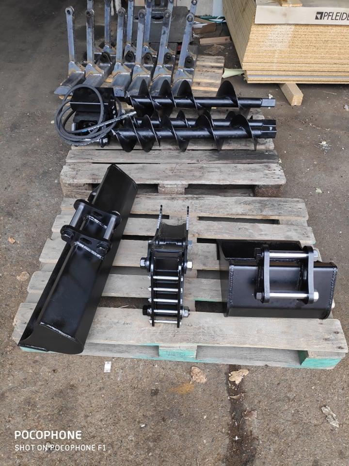 Zestaw czarnych akcesoriów do maszyn budowlanych, w tym wiertła glebowe, łyżki i adapter na palecie w magazynie.