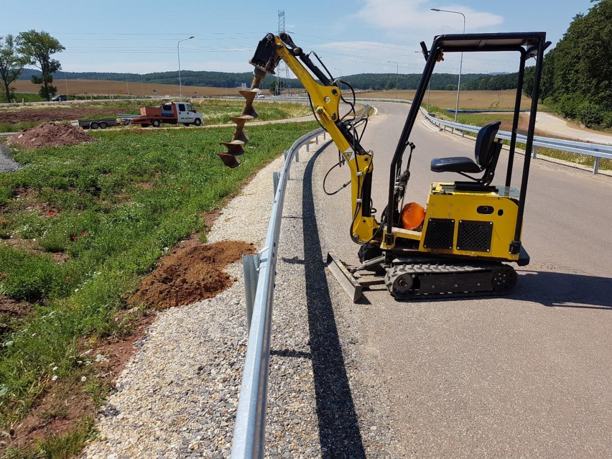 Żółta maszyna z wiertłem wiercąca dziury w ziemi obok barierek na poboczu drogi, w tle widoczny krajobraz z transportem.