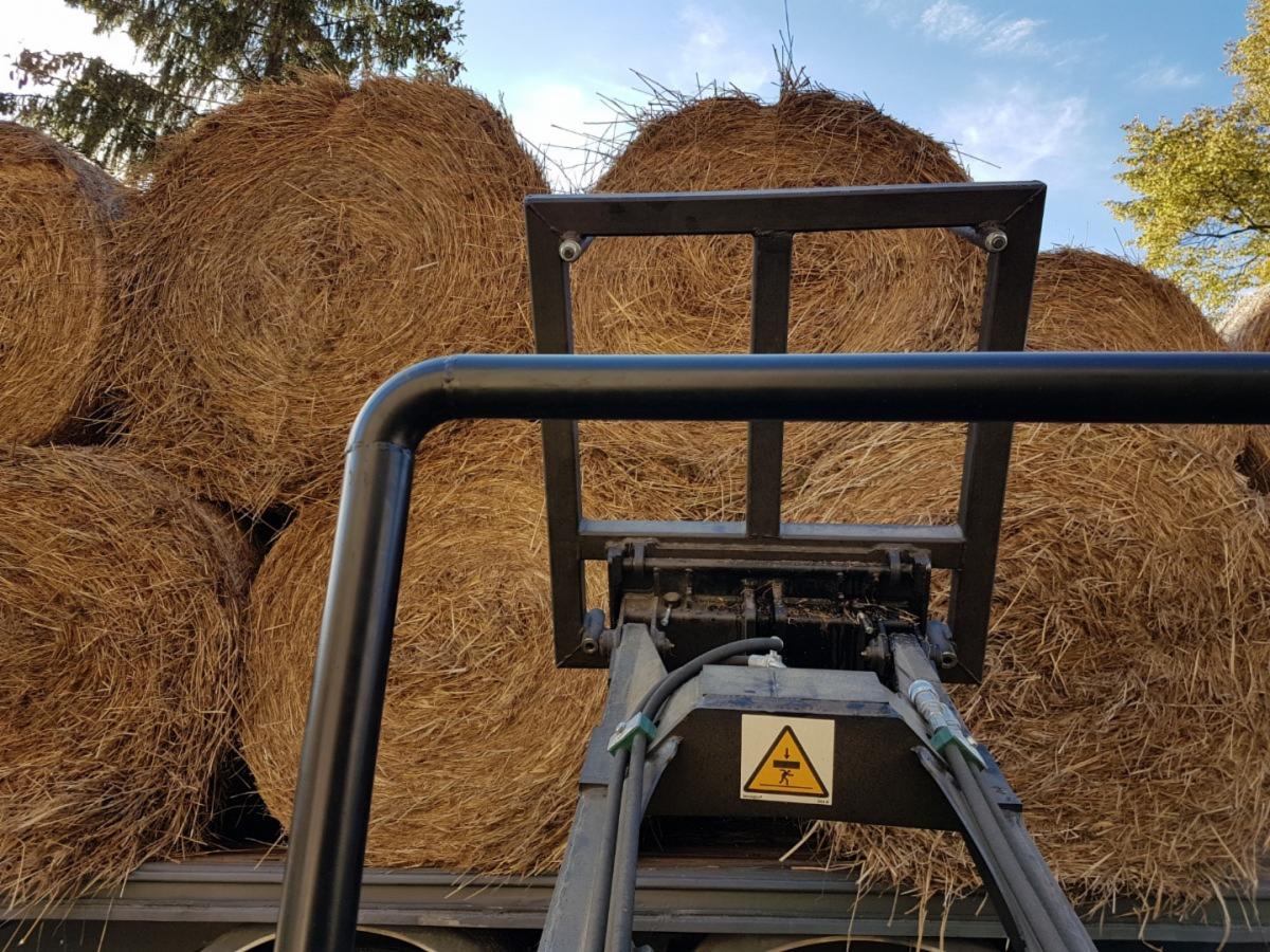 Załadunek okrągłych bel słomy za pomocą osprzętu na maszynie rolniczej, widoczny znak ostrzegawczy.
