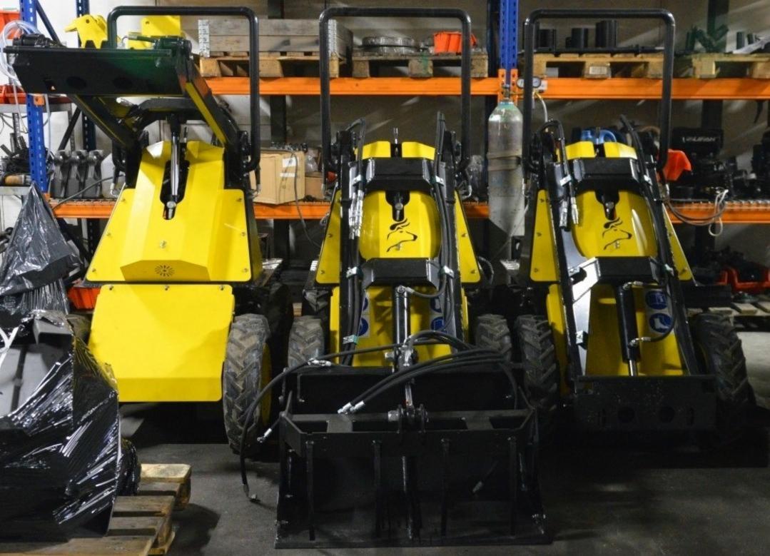 Trzy żółte maszyny budowlane z czarnymi elementami i łyżkami na tle regałów magazynowych; środkowa maszyna z logo zwierzęcia.