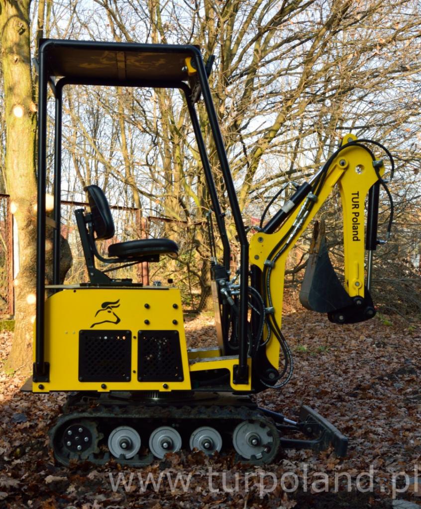Żółta miniładowarka z czarnym ramieniem kopiącym, z logo firmy TUR Poland, na tle drzew bez liści i ziemi pokrytej liśćmi.