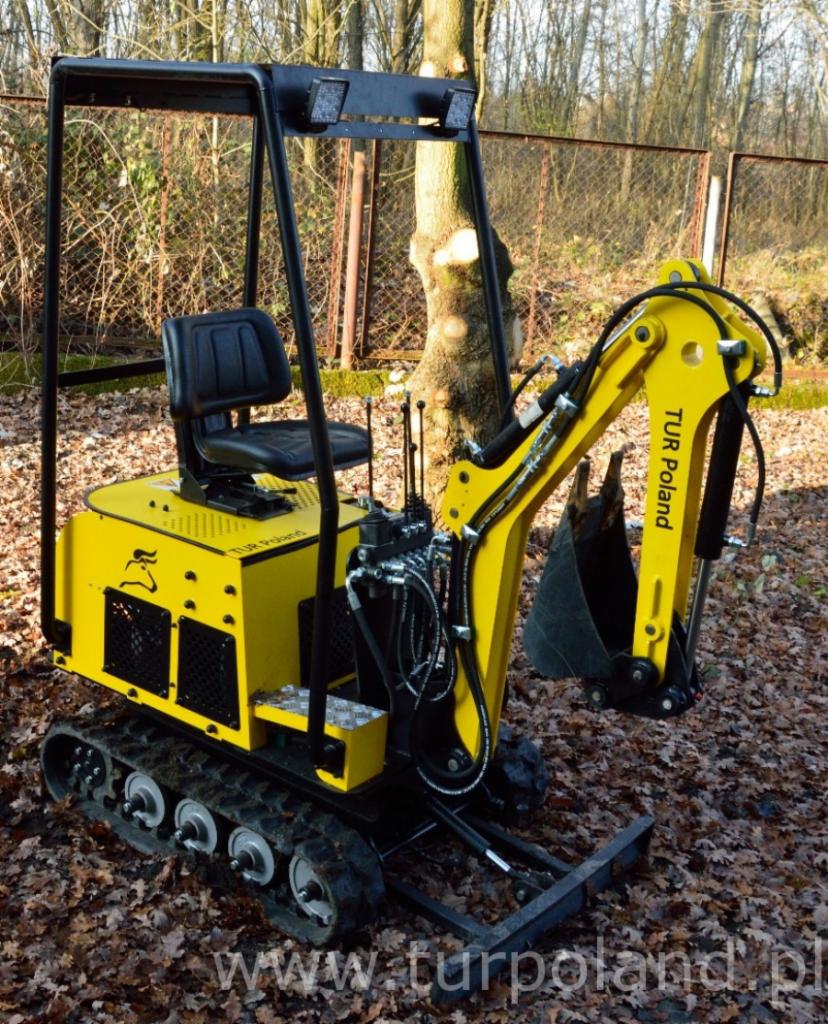 Żółta, gąsienicowa maszyna budowlana z łyżką, marki TUR Poland, zaparkowana na tle drzew i liści