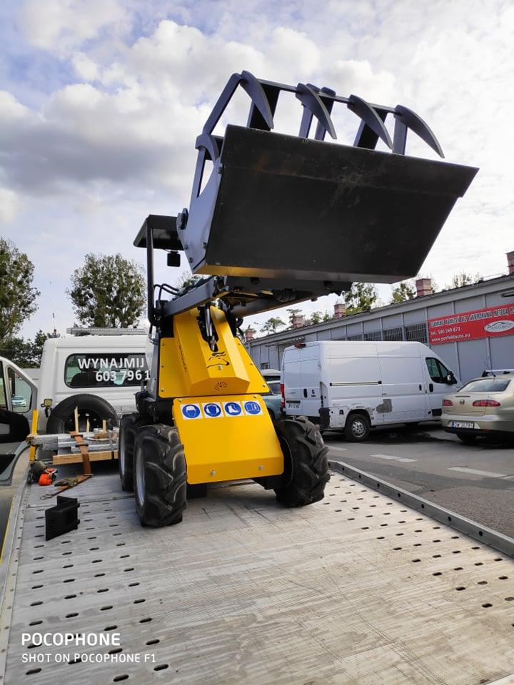 Żółta maszyna budowlana z łyżką na platformie transportowej, widoczne tabliczki informacyjne i numer telefonu do wynajmu.