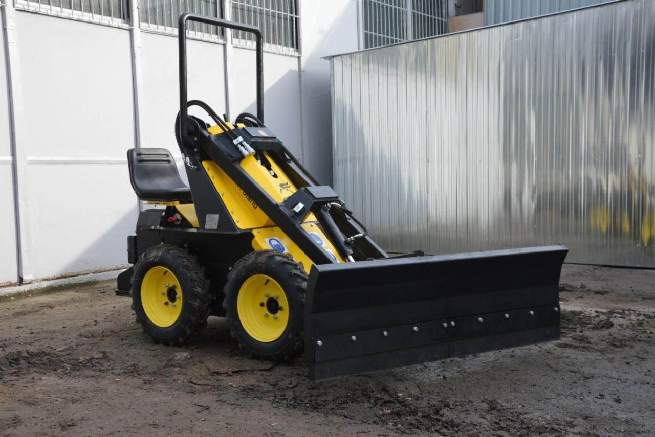 Żółto-czarna maszyna z czarnym pługiem na ziemnym podłożu, metalowe budynki w tle.