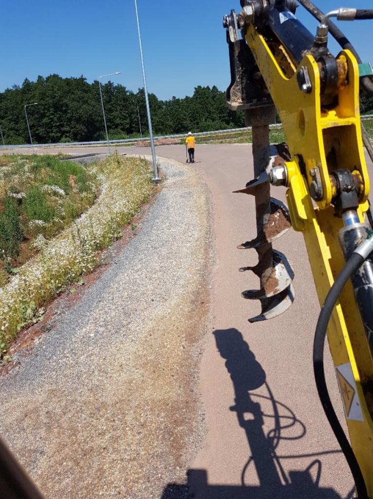 Żółta miniładowarka z wiertłem ziemnym w trakcie pracy na poboczu drogi, w tle pracownik w pomarańczowej kamizelce i zalesiony teren.
