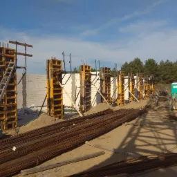 Budowa domu murowanego: szalunki z ż&oacute;łtymi elementami, zbrojenie w wiązkach na piasku i białe bloczki konstrukcyjne, widok na plac budowy z przenośną toaletą w tle.