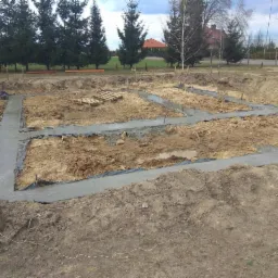 Wylane fundamenty pod budynek z widocznymi ławami fundamentowymi, zabezpieczone folią, na tle wykopu z ziemią i drzewami iglastymi w oddali.