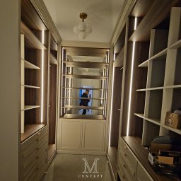 TOTAL RENOVATIONS SPÓŁKA Z OGRANICZONĄ ODPOWIEDZIALNOŚCIĄ - Garderoba w stylu klasycznym z frezowanymi frontami kasetonowymi w kolorze ciemnego orzecha, oświetlona ledowymi listwami. Widoczny fragment osoby w lustrzanym odbiciu.