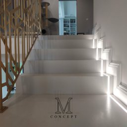 TOTAL RENOVATIONS SPÓŁKA Z OGRANICZONĄ ODPOWIEDZIALNOŚCIĄ - Białe schody z podświetleniem LED i złotą, geometryczną balustradą. W tle widoczny regał z książkami i drapak dla kota. Minimalistyczny design.