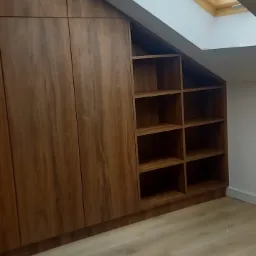 Zabudowa wnęki ze skosem z drewnianymi frontami i otwartymi półkami, oświetlona przez okno dachowe. Podłoga z paneli w odcieniach beżu.