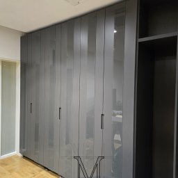 TOTAL RENOVATIONS SPÓŁKA Z OGRANICZONĄ ODPOWIEDZIALNOŚCIĄ - Szara szafa wnękowa z frontami MDF i pasami szkła, zamontowana w pomieszczeniu z parkietem. Widoczny fragment sufitu z klimatyzacją i osoba montująca szafę.