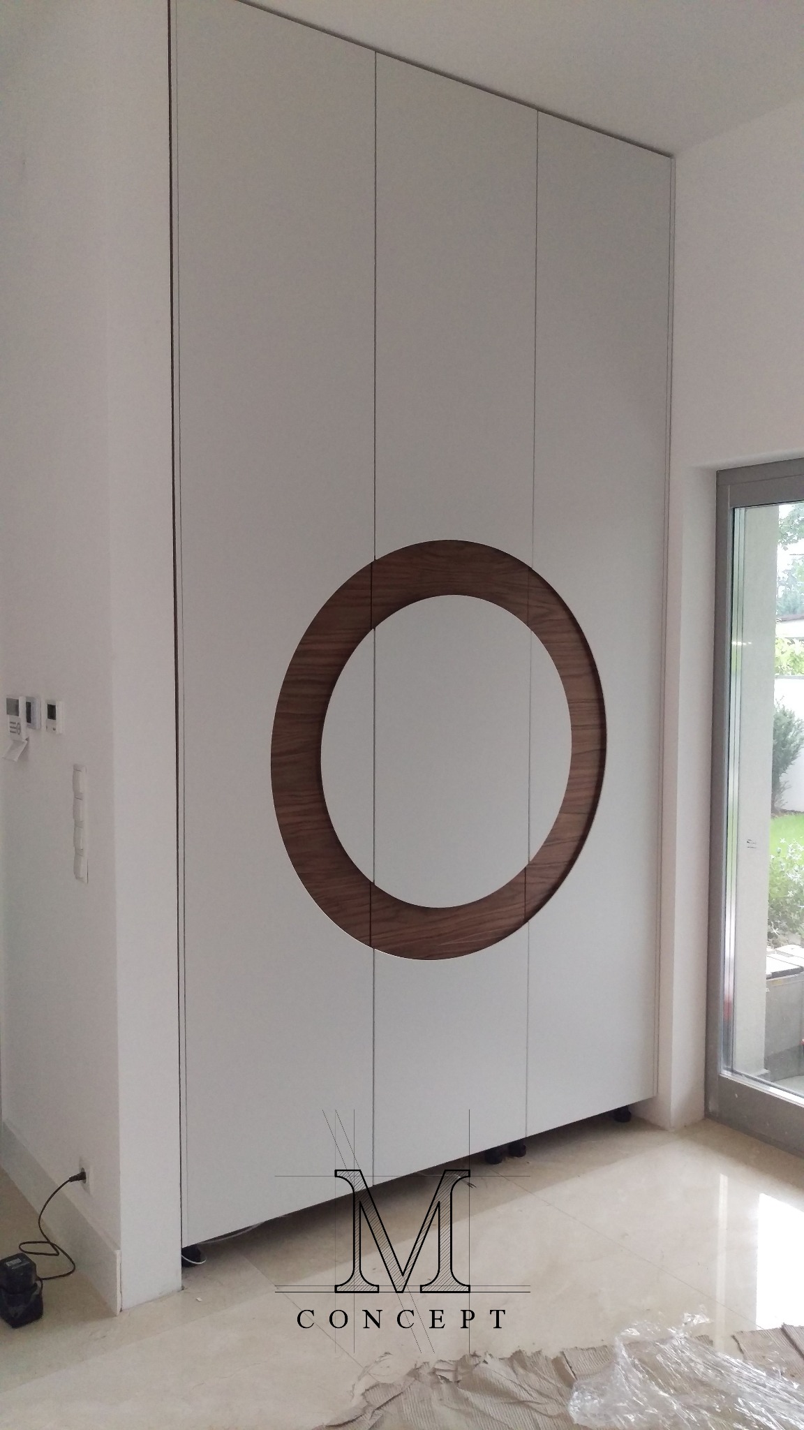 Biała szafa wnękowa z okrągłym, drewnianym elementem dekoracyjnym na środkowych drzwiach, widoczna z boku, w minimalistycznym wnętrzu.