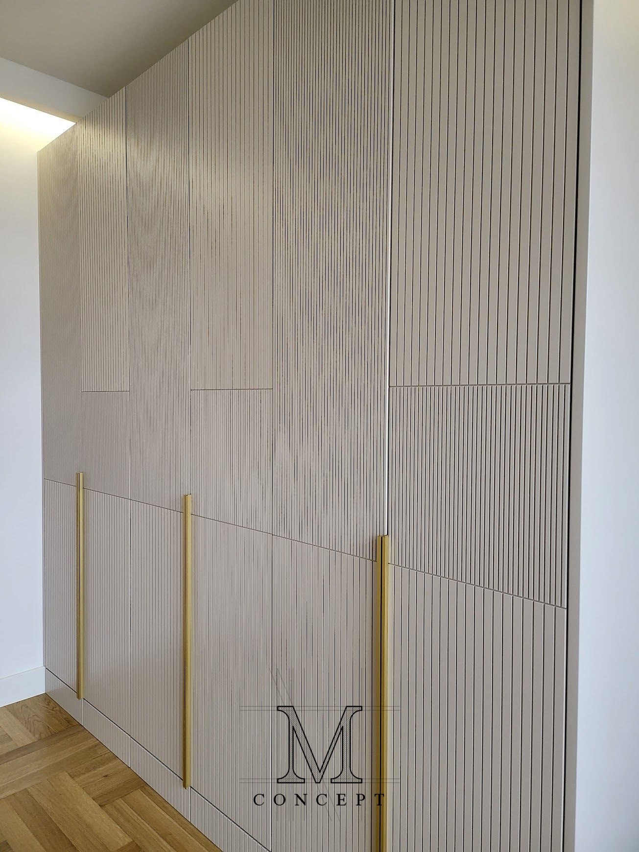 Beżowa szafa wnękowa z ryflowanymi frontami i złotymi uchwytami, widoczna z bliska, z fragmentem parkietu i logo producenta w prawym dolnym rogu.