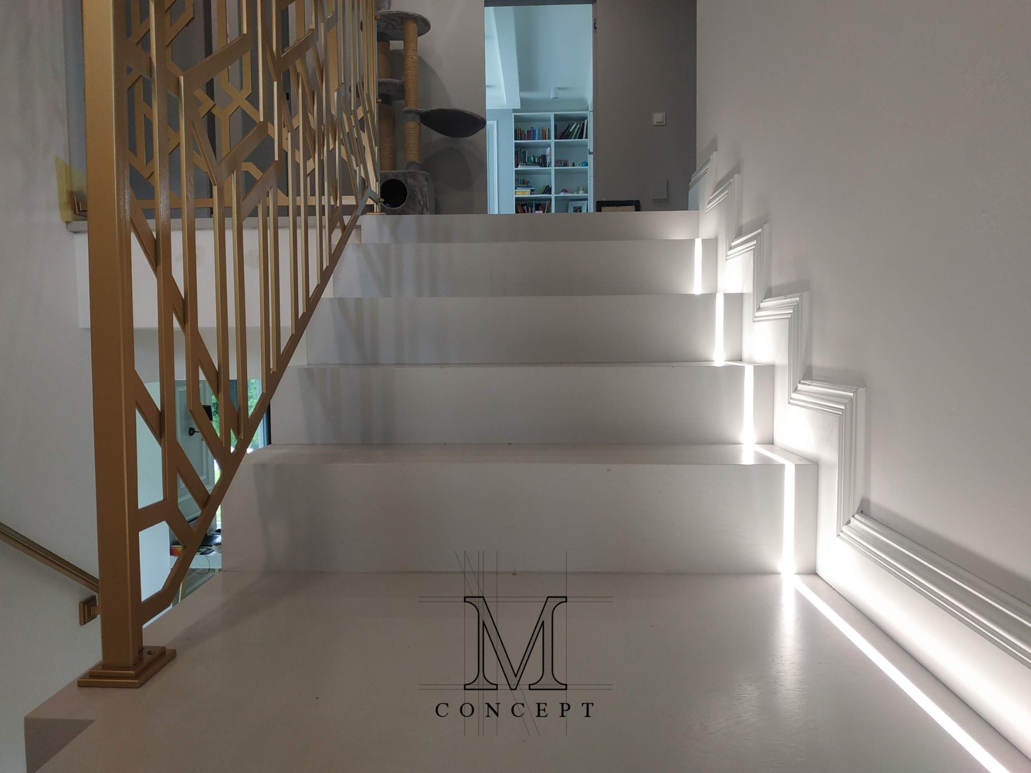 Białe schody z podświetleniem LED i złotą, geometryczną balustradą. W tle widoczny regał z książkami i drapak dla kota. Minimalistyczny design.
