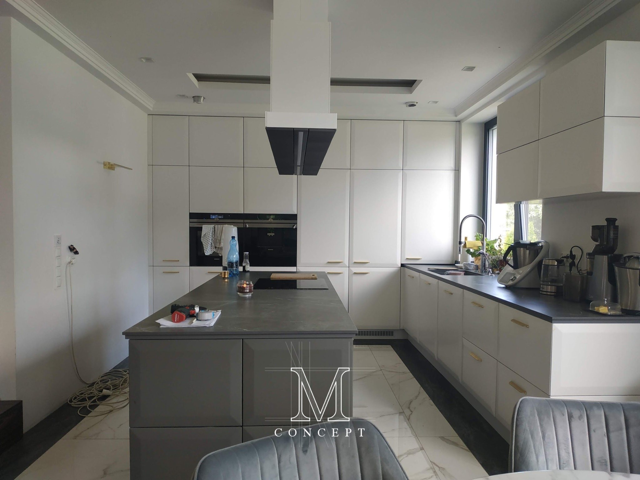 Nowoczesna kuchnia z frezowanymi frontami, szarym blatem Atlas Plan Grey Stone i wyspą. Sprzęt AGD marki Siemens. Widok na marmurową podłogę i szare krzesło.