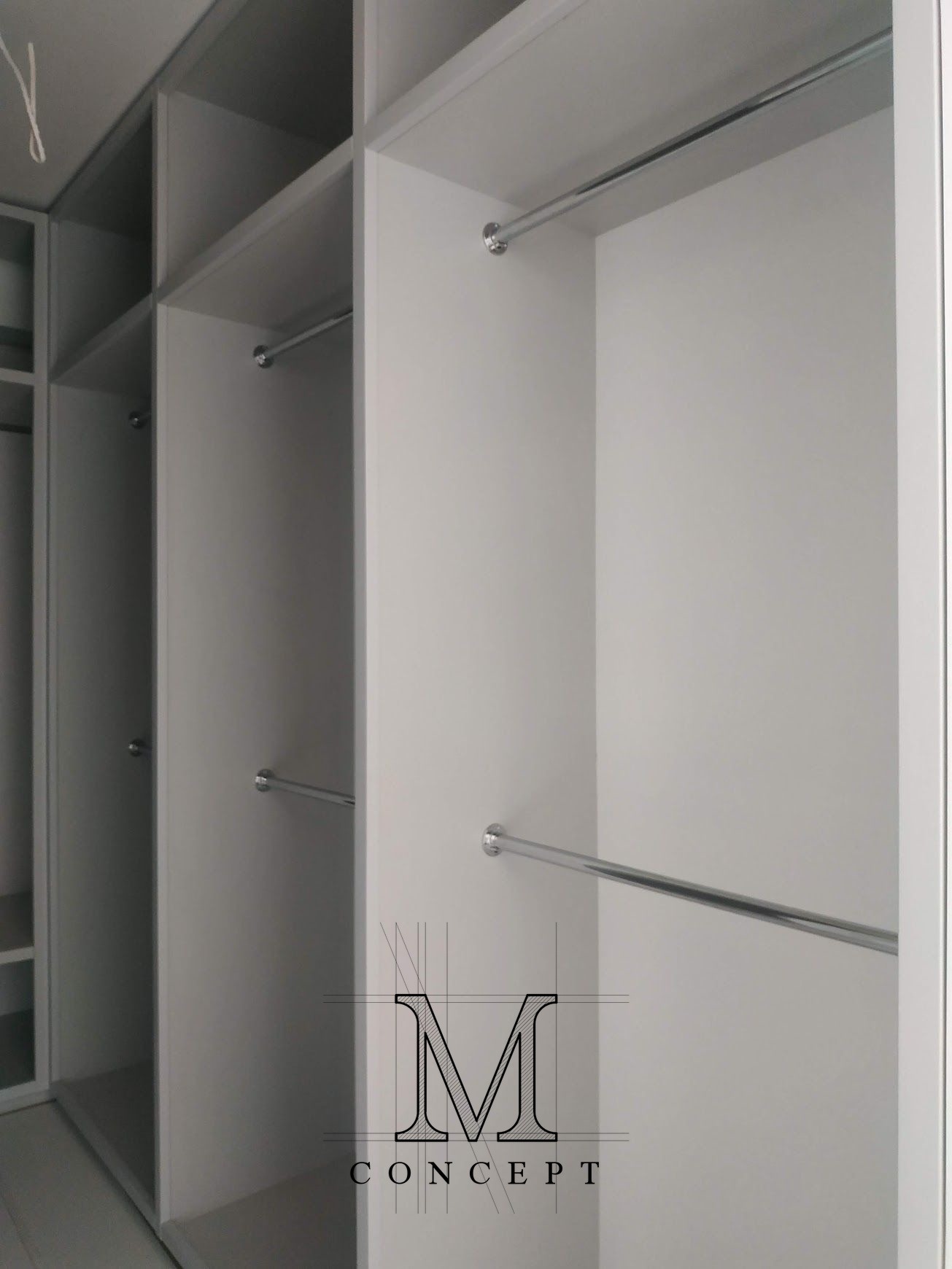 Wnętrze nowoczesnej, białej garderoby z półkami i drążkami na ubrania. Widoczne logo firmy 'M Concept' w prawym dolnym rogu.