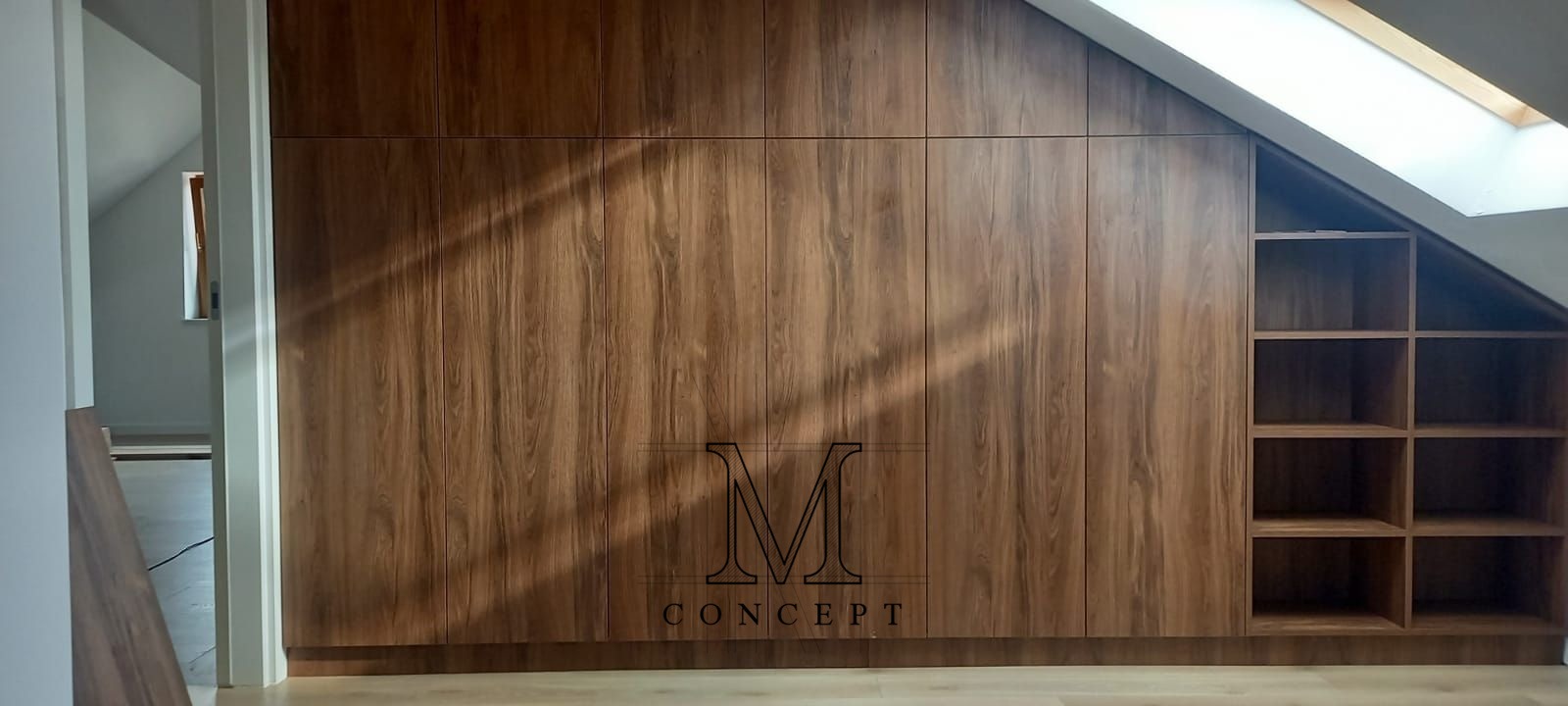 Zabudowa wnęki ze skosem, drewnopodobna szafa z półkami, widok na poddasze z oknem dachowym, minimalistyczny design.