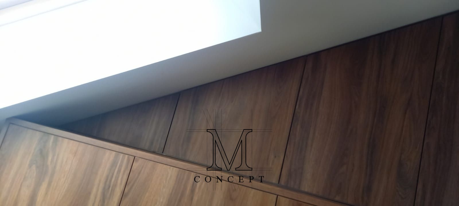 Zabudowa pod skosem z ciemnego drewna, dopasowana do nietypowej przestrzeni. Widoczne panele i górna część zabudowy, oświetlenie sufitowe. Minimalistyczny design.