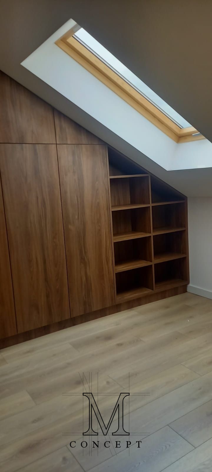 Zabudowa wnęki ze skosem z drewnianymi frontami i otwartymi półkami, oświetlona przez okno dachowe. Podłoga z paneli w odcieniach beżu.