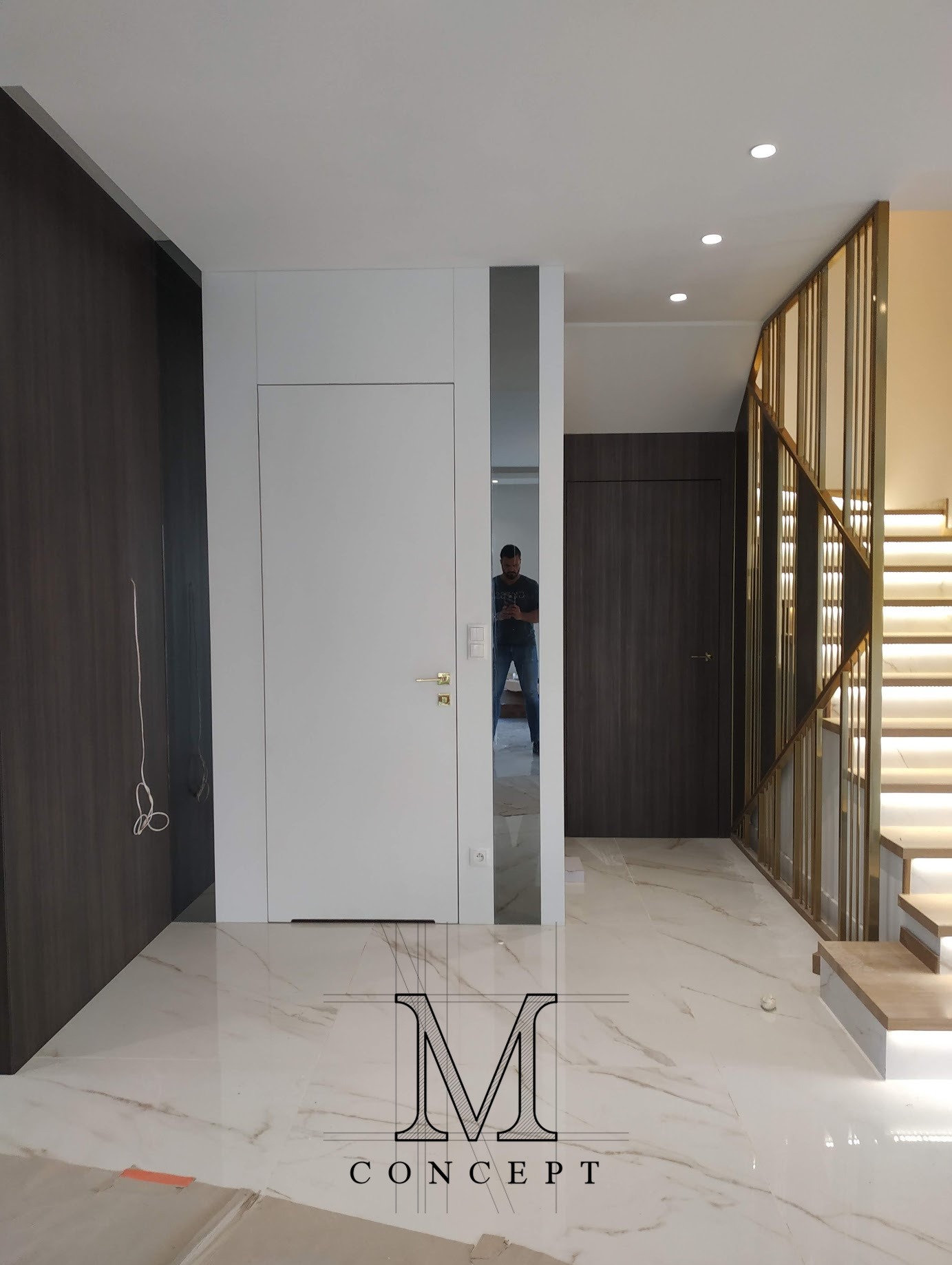 Nowoczesny przedpokój z białymi drzwiami ukrytymi w zabudowie, lustrem i podświetlanymi schodami z metalową balustradą. Minimalistyczny design z marmurową podłogą.