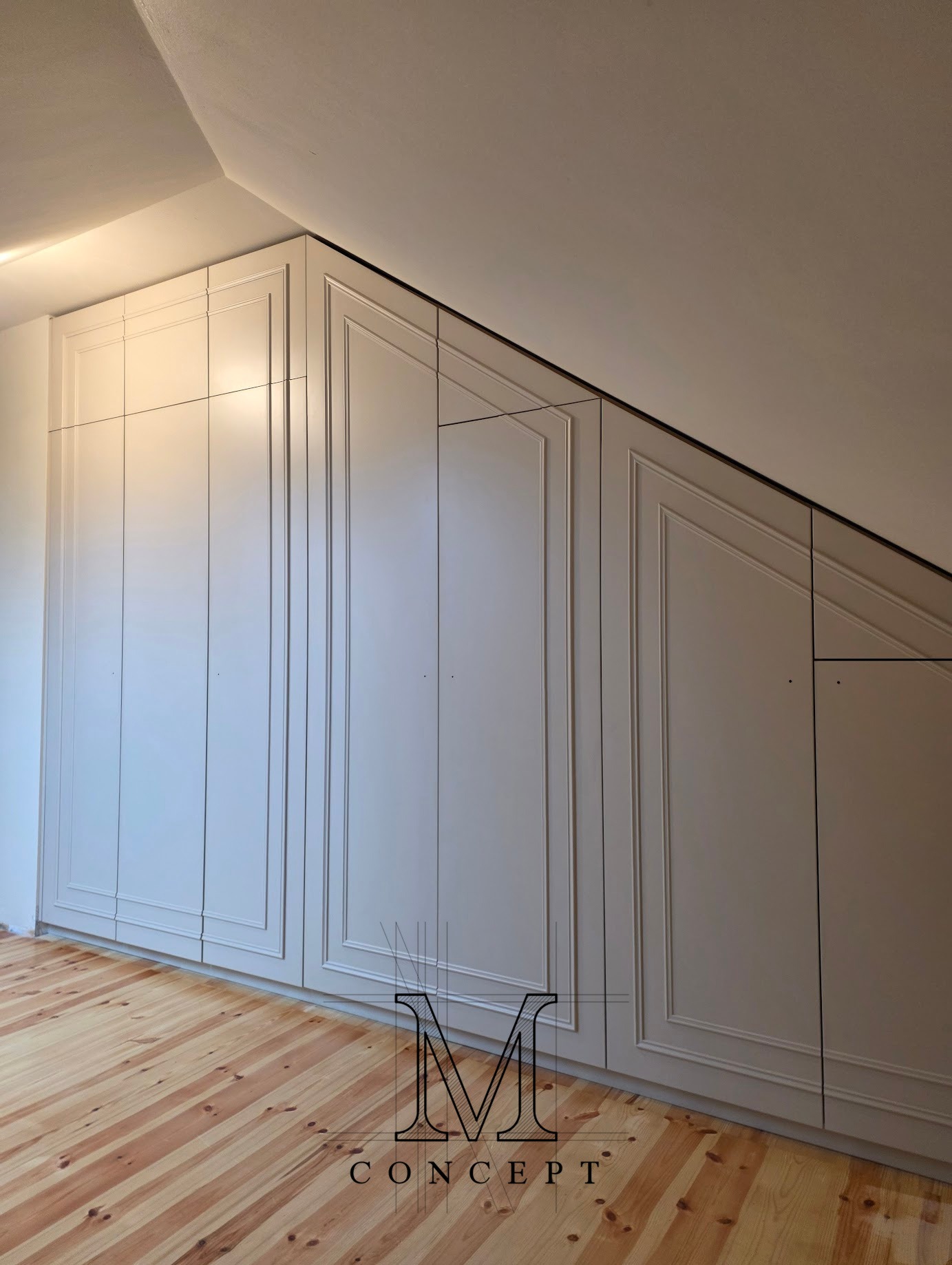 Szafa wnękowa na poddaszu z białymi frontami MDF w stylu klasycznym, dopasowana do skosu dachu. Widoczna drewniana podłoga i oświetlenie.