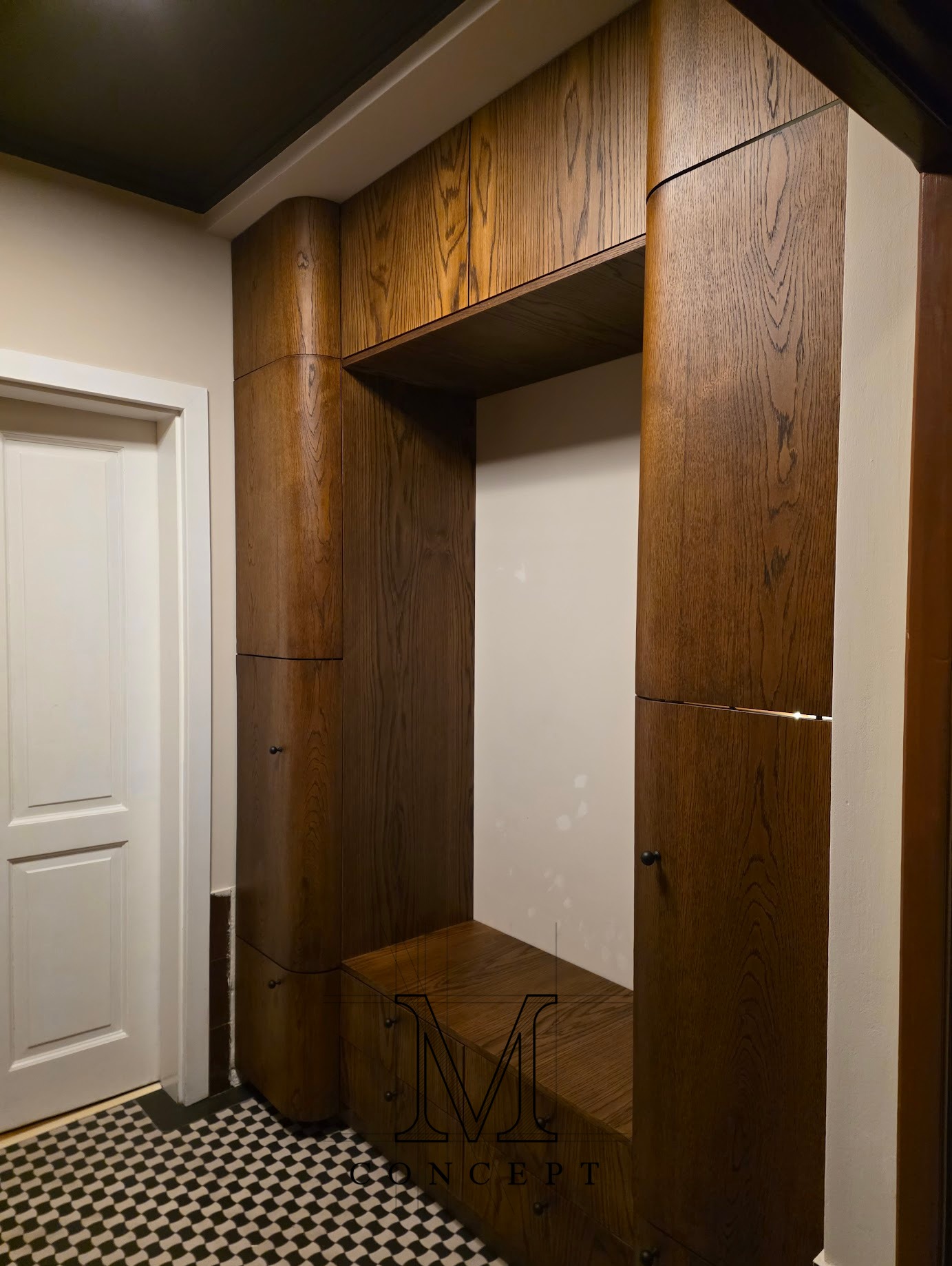Drewniana zabudowa wnęki w przedpokoju z siedziskiem i szafkami, widoczna obok białych drzwi i czarno-białej podłogi. Zaokrąglone krawędzie, ciemne wykończenie drewna.