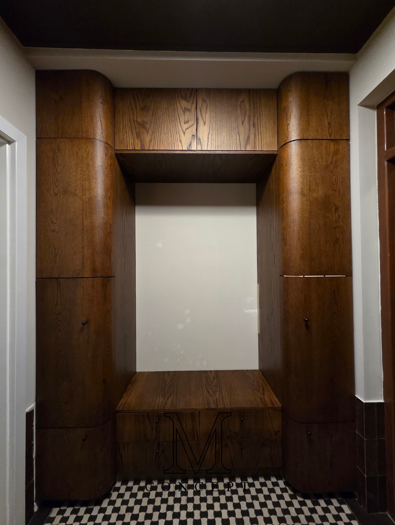 Wnęka z siedziskiem i szafkami z ciemnego drewna o zaokrąglonych krawędziach, na podłodze czarno-biała szachownica. Widok z przodu, oświetlenie górne.