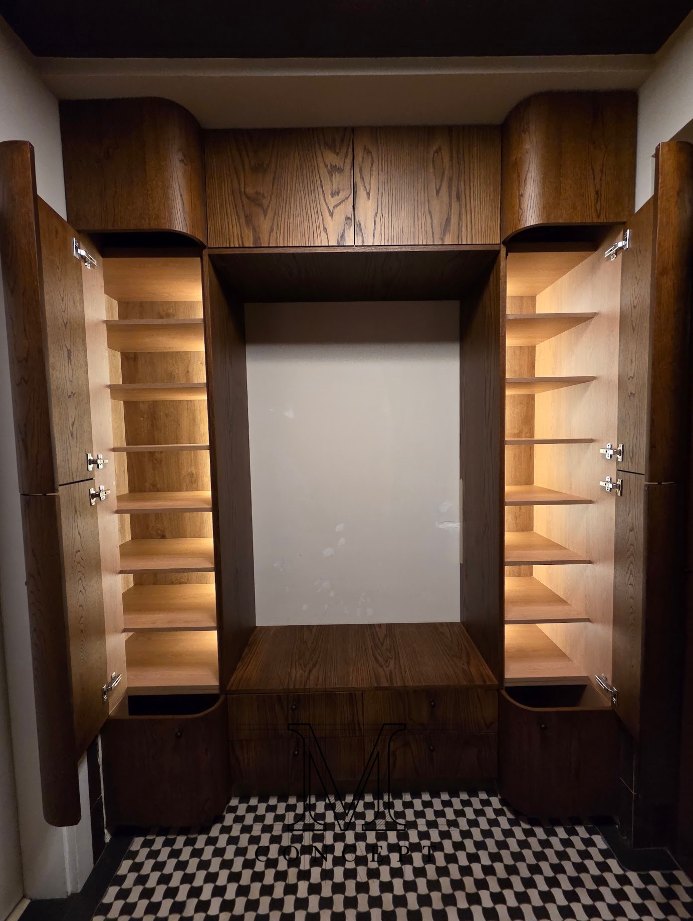 Drewniana szafa wnękowa z oświetleniem wewnętrznym, otwarte drzwi ukazują półki. Na dole szafki z szufladami, podłoga w czarno-białą szachownicę. Ciepłe światło.