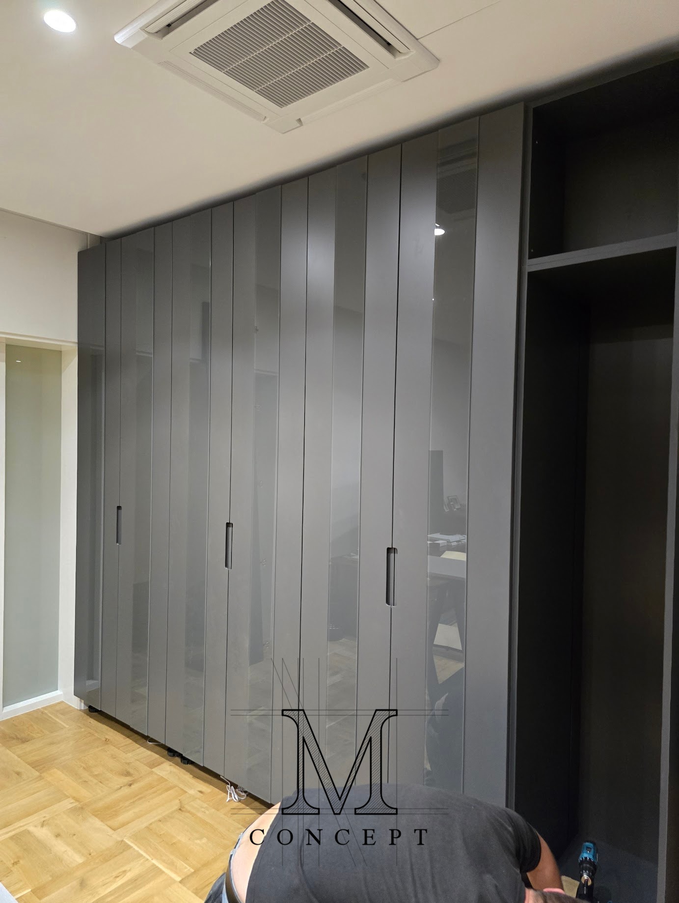 Szara szafa wnękowa z frontami MDF i pasami szkła, zamontowana w pomieszczeniu z parkietem. Widoczny fragment sufitu z klimatyzacją i osoba montująca szafę.