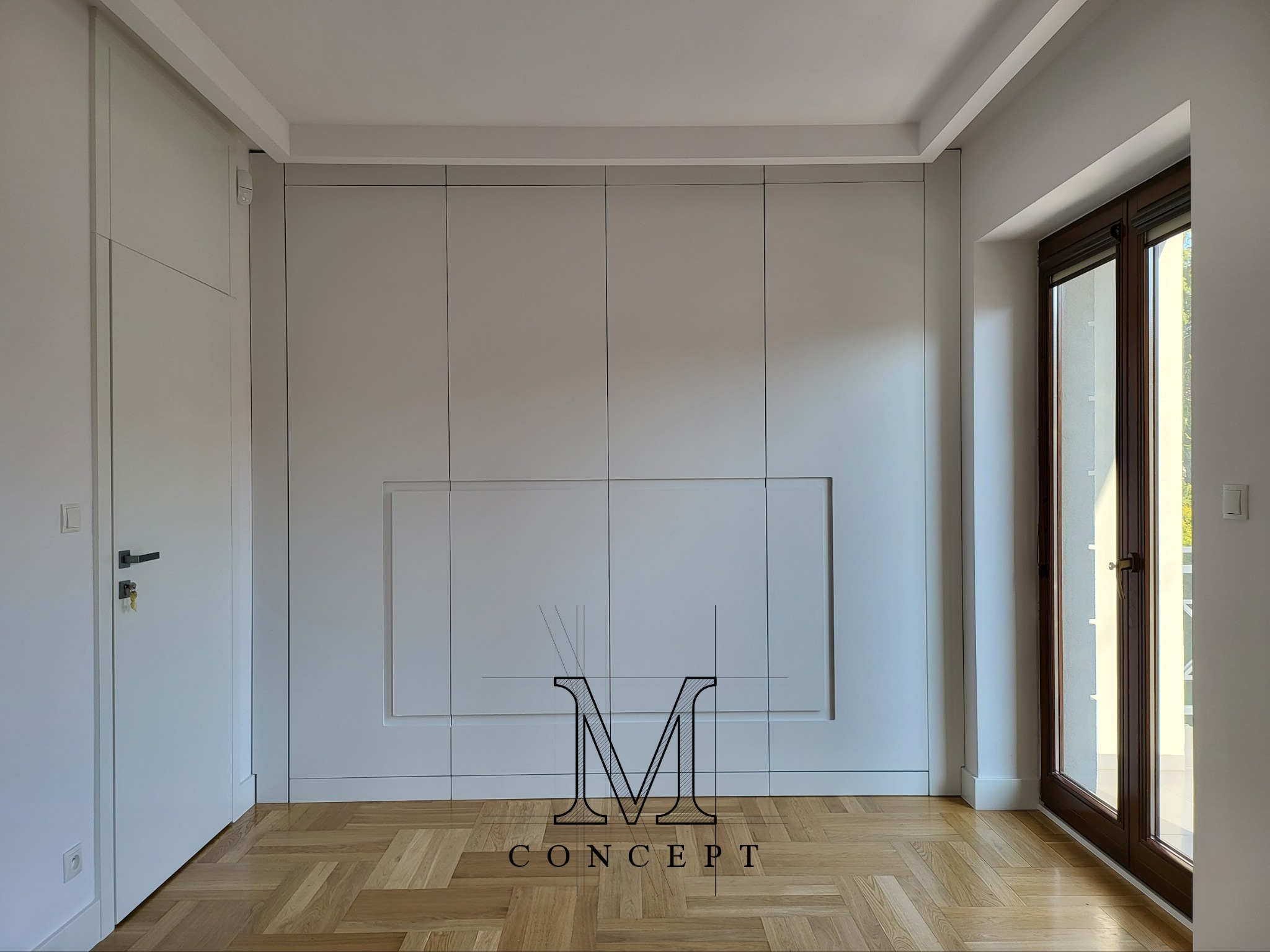 Minimalistyczna szafa wnękowa z frezowanymi uchwytami, lakierowana na półmat, w jasnym odcieniu szarości, z parkietem w jodełkę na podłodze i oknem z ciemną ramą.