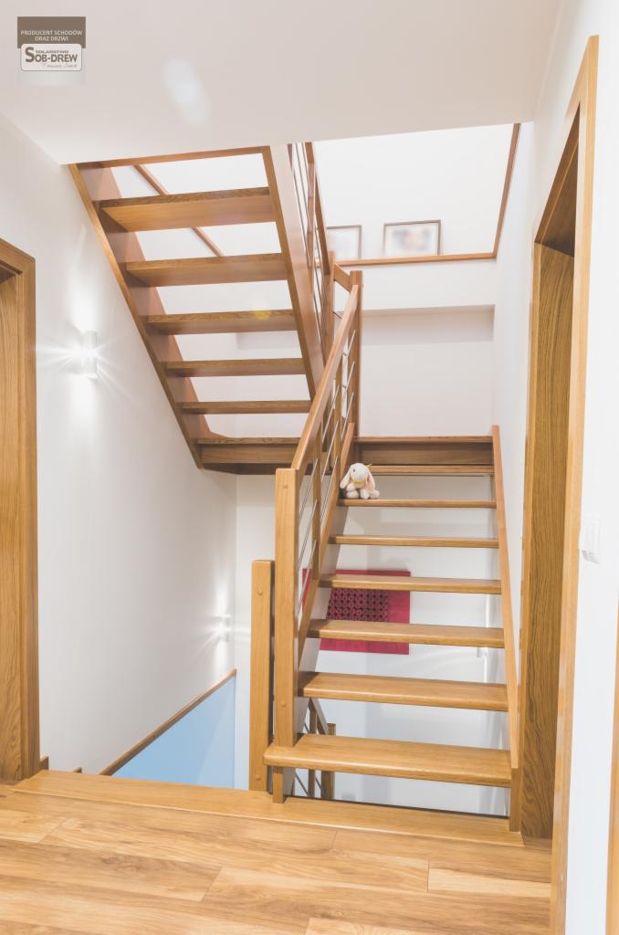 Jasne, dębowe schody policzkowe z relingami ze stali nierdzewnej, prowadzące na wyższą kondygnację. Na jednym ze stopni pluszowy królik. Widoczne drzwi z ościeżnicą z tego samego drewna.