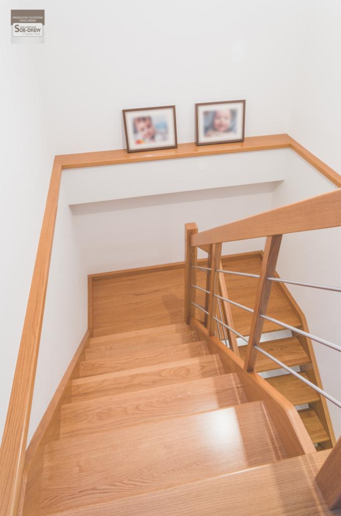 Dębowe schody policzkowe z balustradą z relingami ze stali nierdzewnej, widok z góry, na ścianie dwa zdjęcia w ramkach.